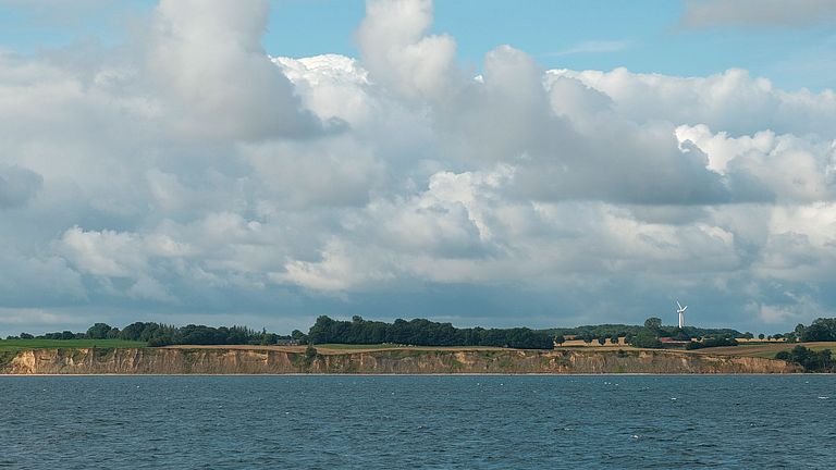 A cliff covered in greenery, the sea in front of it, a blue sky with white clouds above