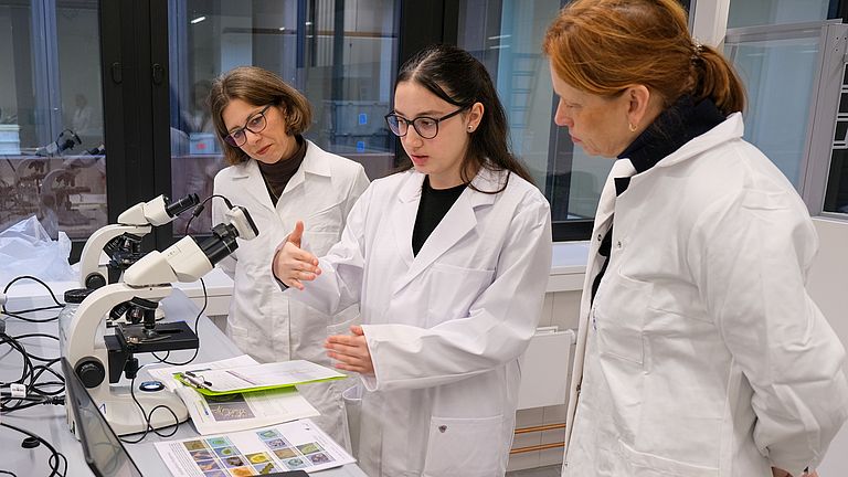 Drei Frauen stehen in einem Labor vor zwei Mikroskopen, einem Laptop und einem aufgeschlagenen Buch