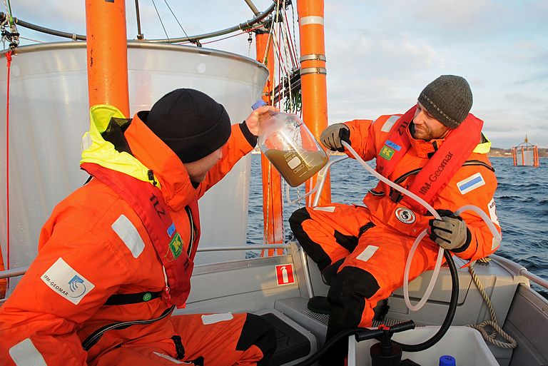 Zwei Forscher in roten Überlebensanzügen arbeiten von einem Boot aus an einem Mesokosmos. 