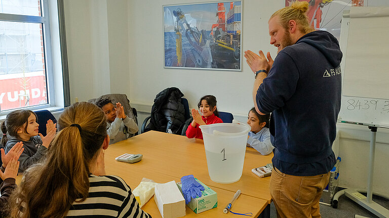 Ein Mann mit blondem Zopf und blauem Hoodie steht an einem Tisch, an dem Schulkinder sitzen und reibt die Hände kräftig aneinander