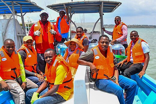 Zehn dunkelhäutige Personen mit lauchtend orangen Schwimmwesten auf einem blauen Boot