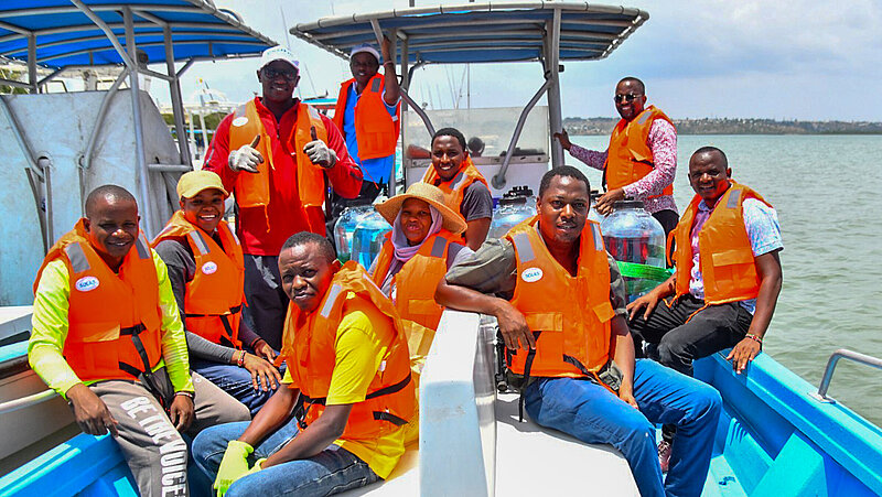 Zehn dunkelhäutige Personen mit lauchtend orangen Schwimmwesten auf einem blauen Boot