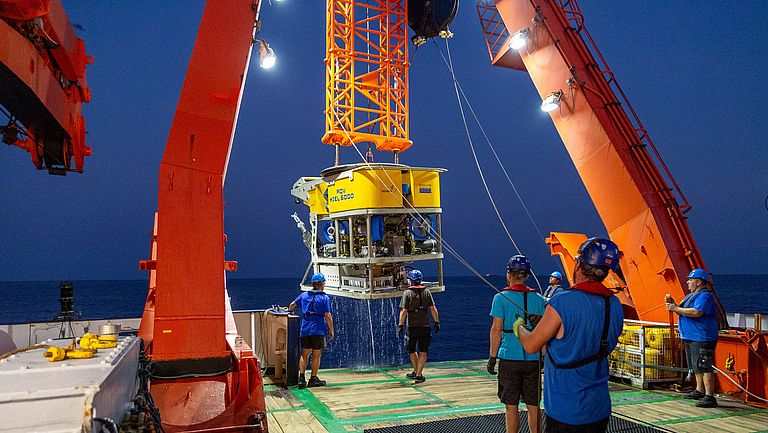 A yellow submersible is lifted out of the water by a red crane on board a research vessel.