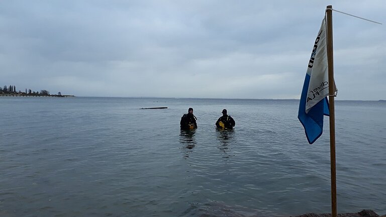 Zwei Taucher stehen an einem Ostseestrand bis zur Hüfte im Wasser. Ihre tauchanzüge sind schwarz. Sie kommen gerade aus dem Wasser und sind mit gelben Taschen beladen. Der Himmel ist bedeckt. Im Vordergrund ist eine Taucherflagge aufgestelt, die schapp herunter hängt, da kein Wind weht. 