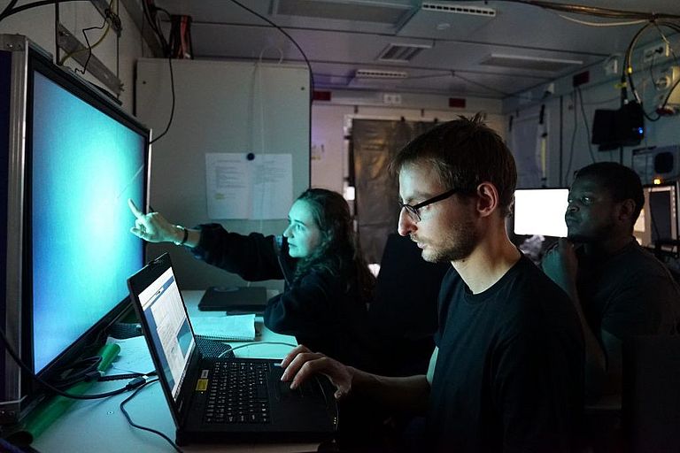 zwei Wissenschaftler und eine Wissenschaftlerin sitzen in einem dunklen Schiffslabor vor leuchtenden Bildschirmen. 
