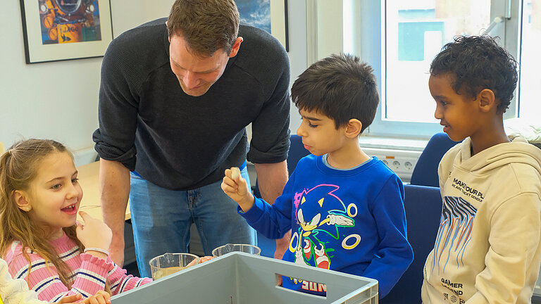 Ein Mann beugt sich über einen Tisch, an dem Schulkinder sitzen und Strandfunde aus einer Wanne betrachten