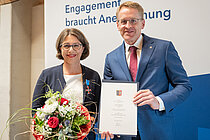 Eine Frau mit Blumenstraß und ein Mann mit Urkunde lächeln in die Kamera