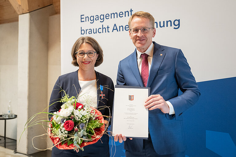 A woman holding a bouquet of flowers and a man holding a certificate are smiling at the camera