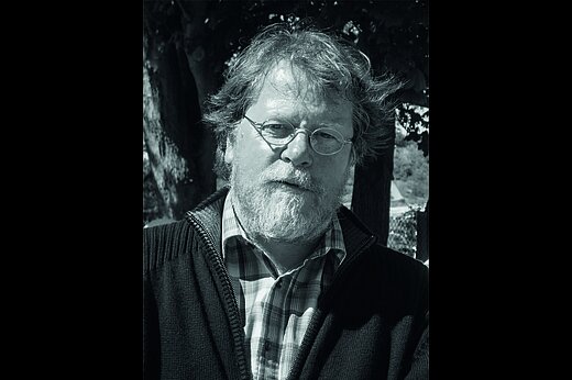 Black-and-white photograph of an elderly man with round glasses, tousled hair and a full beard. He is wearing a checked shirt under a woollen jumper with a zipper