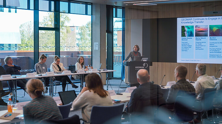 A woman is standing at a lectern in a conference room giving a presentation; the audience is seated at tables