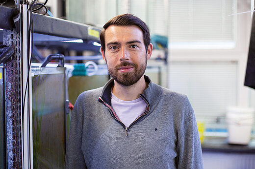 A young man with a dark beard is standing in a research setting with silver-coloured tanks