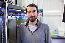 A young man with a dark beard is standing in a research setting with silver-coloured tanks