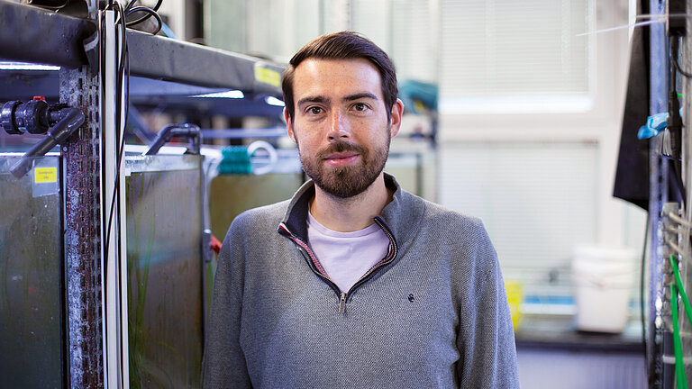 A young man with a dark beard is standing in a research setting with silver-coloured tanks