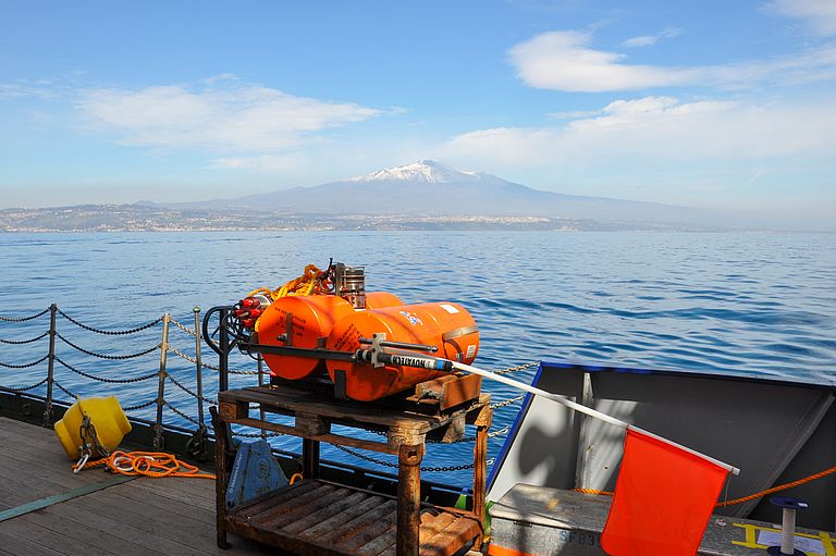 Wissenschaftliche Messgeräte an Bord eines Schiffes, im Hintergrund die Küste mit einem Vulkankegel