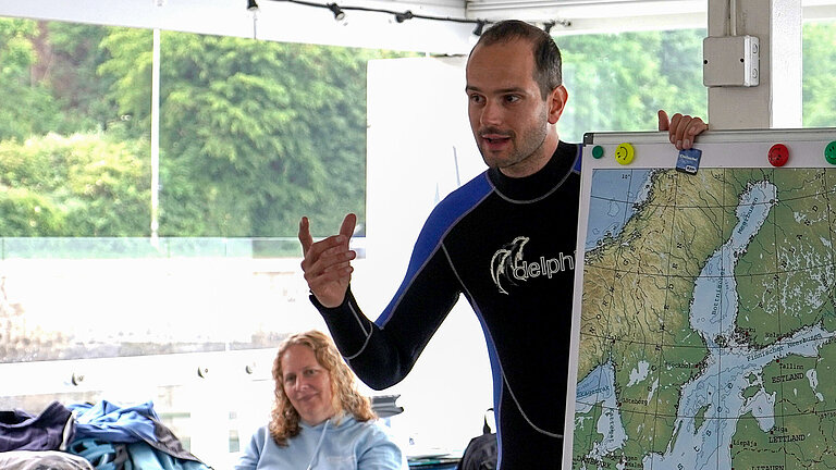 A young man in a wetsuit is standing behind a board displaying a picture of the Baltic Sea. He raises his hand to explain.