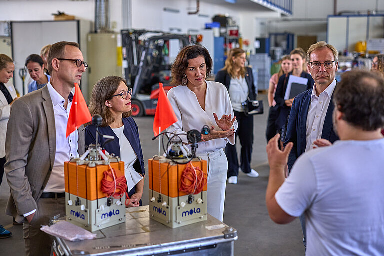 A group of formally dressed people stand in a semicircle listening to a scientist in a white T-shirt, with small orange measuring devices in the foreground.