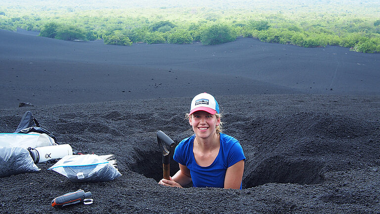 A young woman with a spade peers out of a hole in the black lava sand