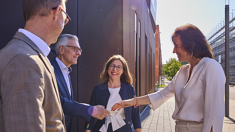 Zwei Männer im Anzug und zwei Frauen im Hosenanzuge begrüßen sich vor einem rostfarbenen Gebäude