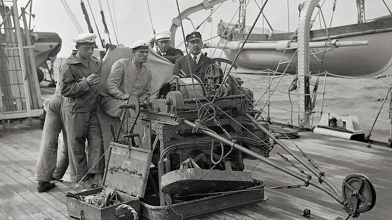 Schwarz-weiß-Fotografie von Seeleuten an Deck, die an einem Holzkasten mit Rädern und Seilen stehen