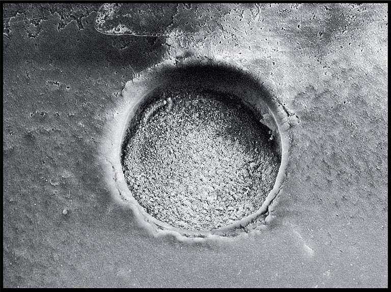 A high-resolution image taken under a scanning electron microscope of an impact crater in volcanic glass. A circular depression can be seen