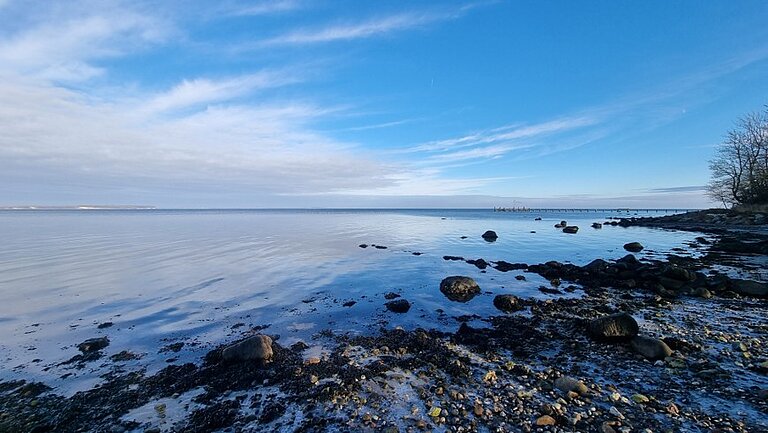 Ein steiniger Ostseestrand im Abendlicht. Das Wasser ist spiegelglatt. Der Himmel im Vordergrund blau, im Hintergrund sind weiße Wolken.