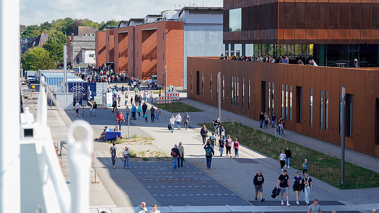 A view from above of a street with passers-by; on the roof terrace of a rust-red building, there are lots of people standing