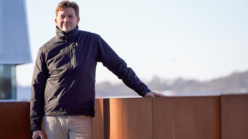 Ein Mann steht an eine rostroten Balustrade, im Hintergrund in der Ferne die Silhouette einer Stadt