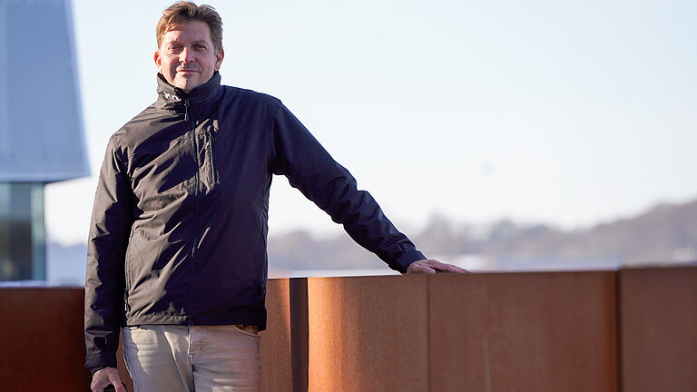 Ein Mann steht an eine rostroten Balustrade, im Hintergrund in der Ferne die Silhouette einer Stadt
