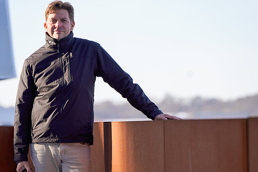 A man stands by a rust-red balustrade, the silhouette of a city visible in the distance behind him.