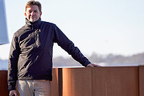 Ein Mann steht an eine rostroten Balustrade, im Hintergrund in der Ferne die Silhouette einer Stadt