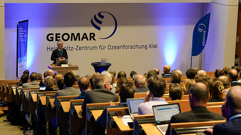 Ein Mann spricht in einem gut gefüllten Hörsaal, im Hintergrund ist der Schriftzug "GEOMAR Helmholtz-Zentrum für Ozeanforschung Kiel" zu erkennen.
