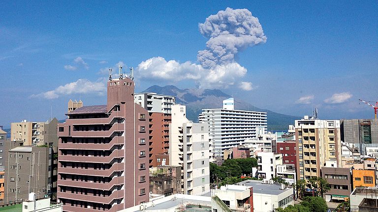 Blick auf den rauchenden Sakurajima mit der Skyline der japanischen Stadt Kagoshima. 