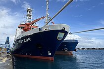 A ship is moored to the harbour wall with ropes. The sky is blue. 