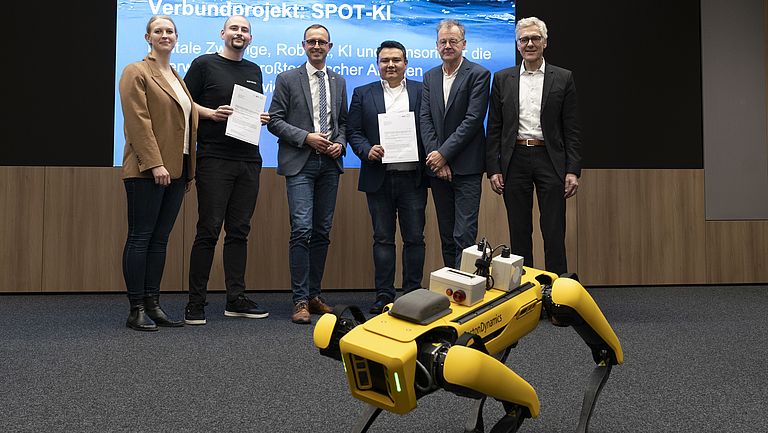 Sechs Menschen stehen in einem Konferenzraum, im Vordergrund ein gelber Roboterhund