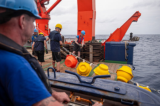 Männer mit Schutzhelmen an Bord eines Schiffes ziehen eine Kette mit gelben und roten Auftriebskörpern aus dem Wasser