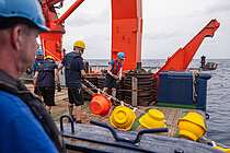 Men wearing hard hats on board a ship are pulling a chain of yellow and red buoys out of the water