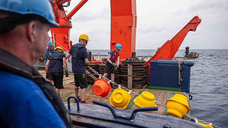 Männer mit Schutzhelmen an Bord eines Schiffes ziehen eine Kette mit gelben und roten Auftriebskörpern aus dem Wasser