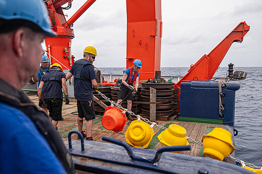 Männer mit Schutzhelmen an Bord eines Schiffes ziehen eine Kette mit gelben und roten Auftriebskörpern aus dem Wasser