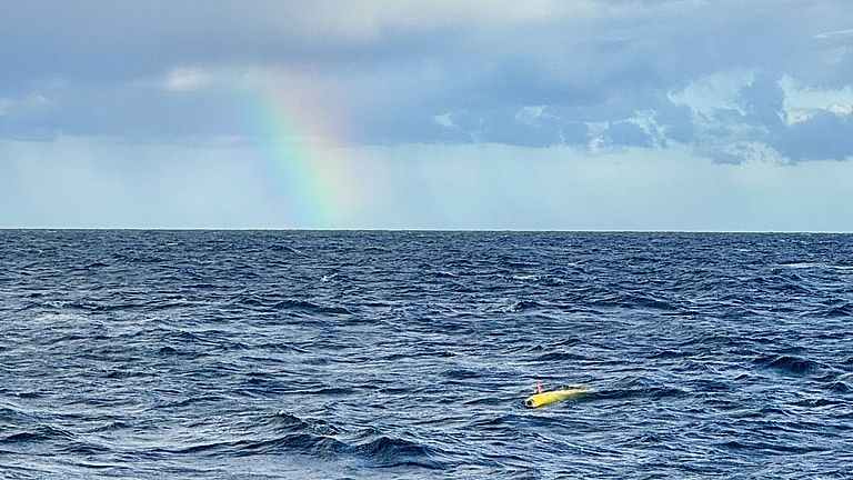 Blick auf das Meer, wo ein kleines gelbes torpedoförmiges Gerät schwimmt, darüber ein Regenbogen