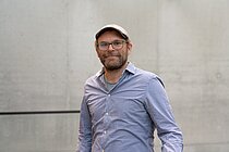 A man with three-day stubble, wearing a flat-brimmed cap, a light blue shirt and glasses, smiles at the camera