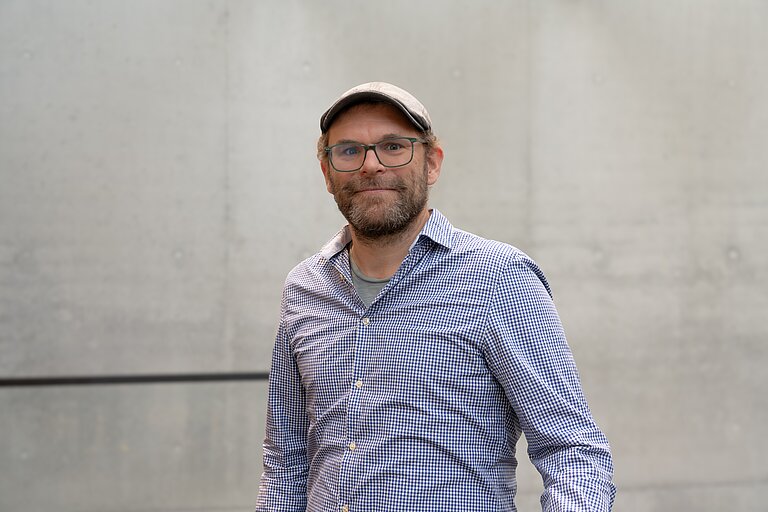 A man with three-day stubble, wearing a flat-brimmed cap, a light blue shirt and glasses, smiles at the camera