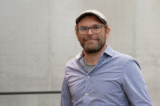Ein Mann mit Drei-Tage-Bart und Schiebermütze und hellblauem Hemd und Brille lächelt in die Kamera