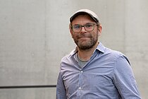 Ein Mann mit Drei-Tage-Bart und Schiebermütze und hellblauem Hemd und Brille lächelt in die Kamera