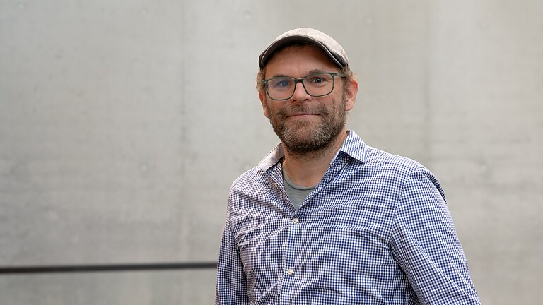 Ein Mann mit Drei-Tage-Bart und Schiebermütze und hellblauem Hemd und Brille lächelt in die Kamera