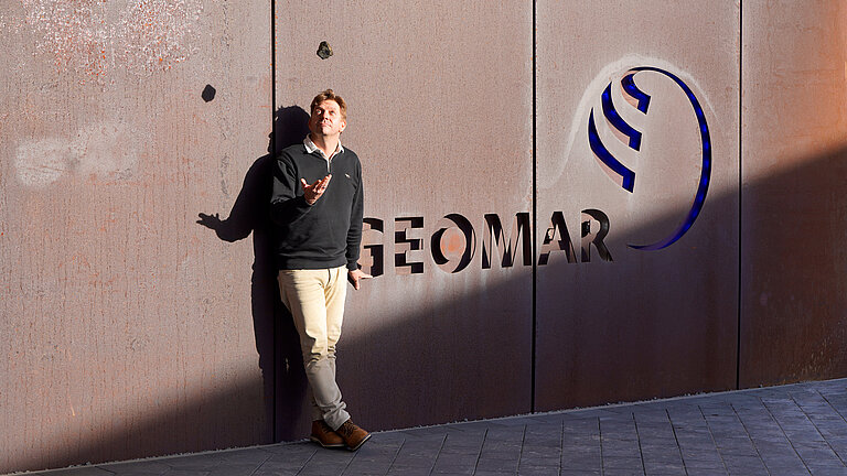 Ein Mann steht vor einer rostroten Fassade, die das Logo "GEOMAR" trägt und wirft einen Stein in die Luft
