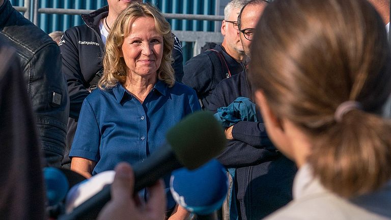 Eine blonde Frau steht inmitten von Pressevertreter:innen mit Mikrofonen