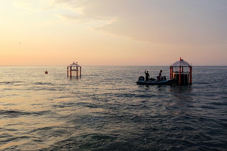 Zwei Mesokosmen und ein Arbeitsboot im Meer vor einem rötlichen Horizont.