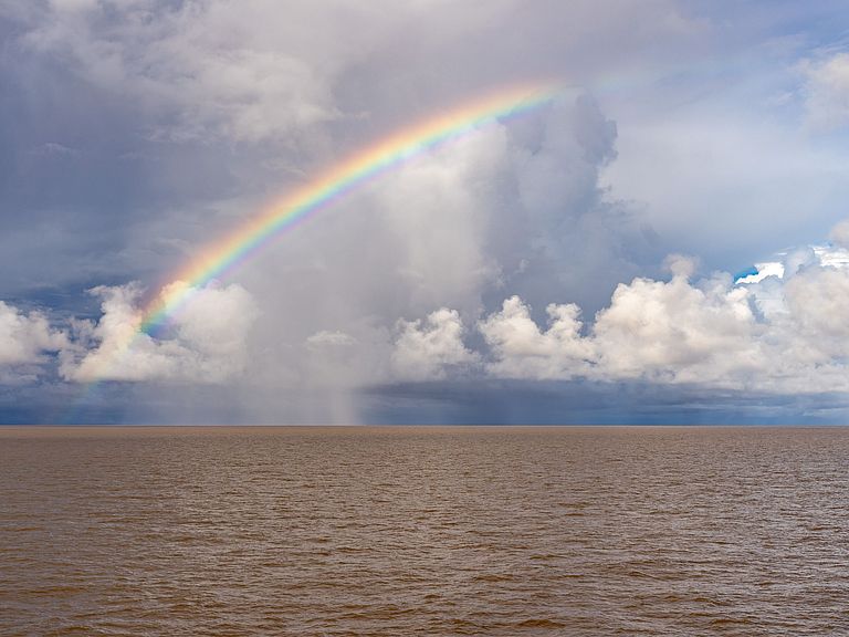 Ein halber Regenbogen überspannt den Himmel über einem schmutzig-braunen Meer.