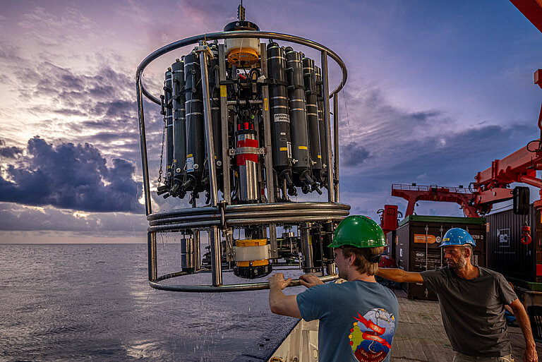 A man wearing a green helmet carries a scientific device from the ship into the water.