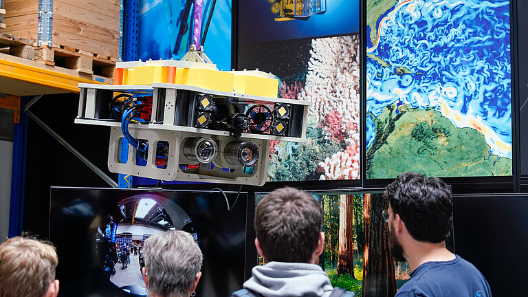 An underwater robot fitted with cameras is suspended in a hall, with visitors standing in front of it, looking on with interest